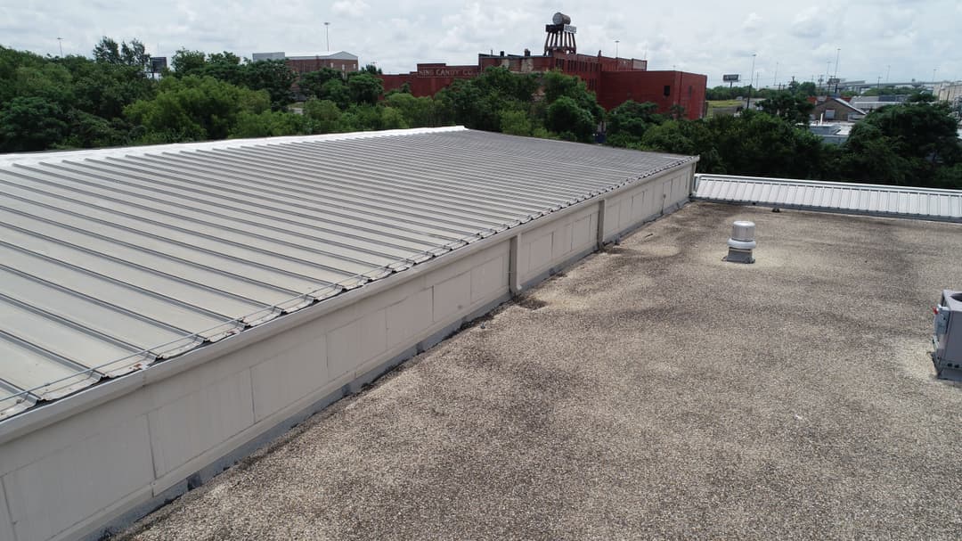 Aerial view of a flat commercial roof with gravel and a ventilation unit, city buildings in background.