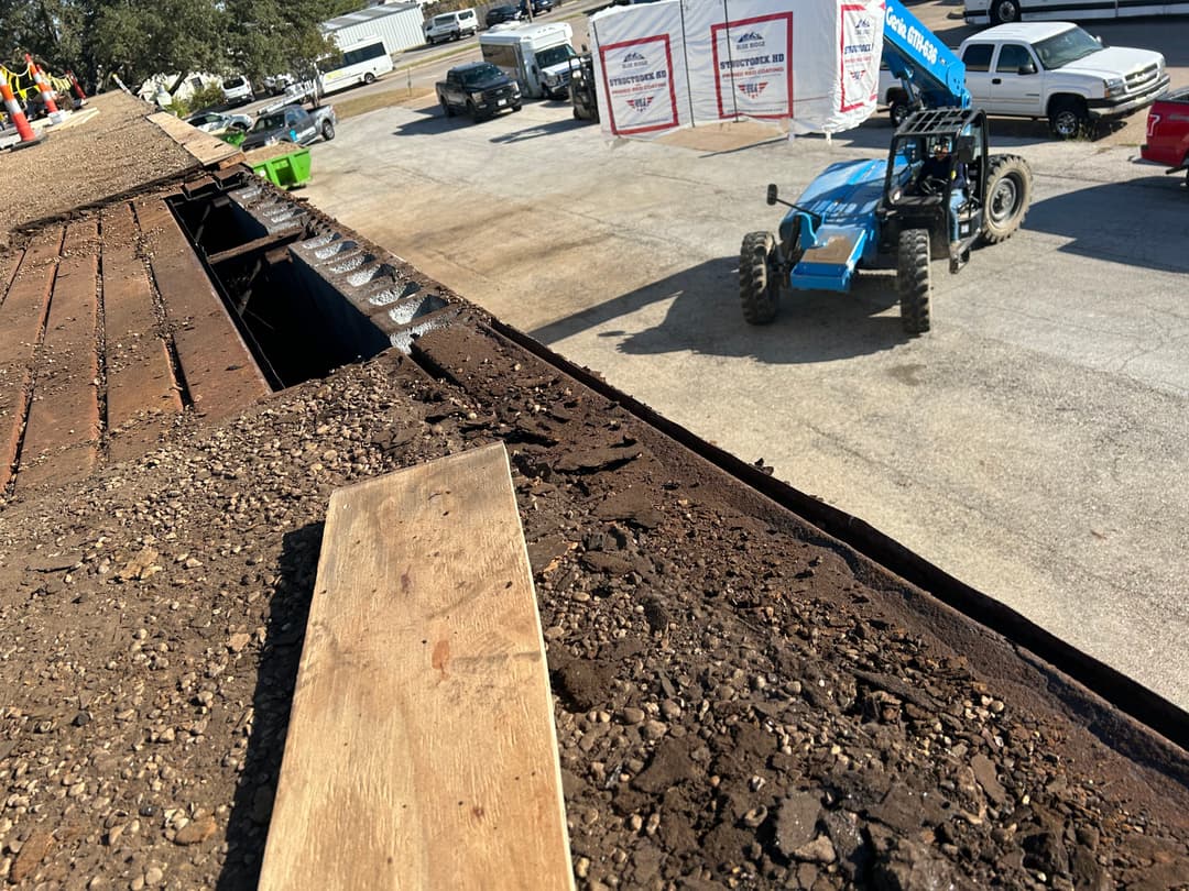 Construction site with heavy machinery, dirt-covered roof edge, and nearby vehicles.