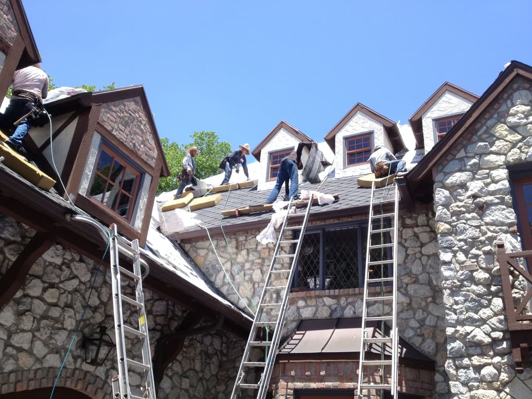 Roofing crew working on a stone house under a clear blue sky with ladders and materials.