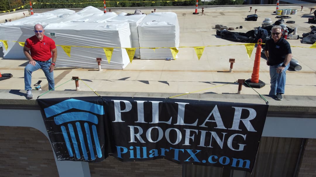 Two men on a rooftop beside a large banner for Pillar Roofing, with construction materials in background.
