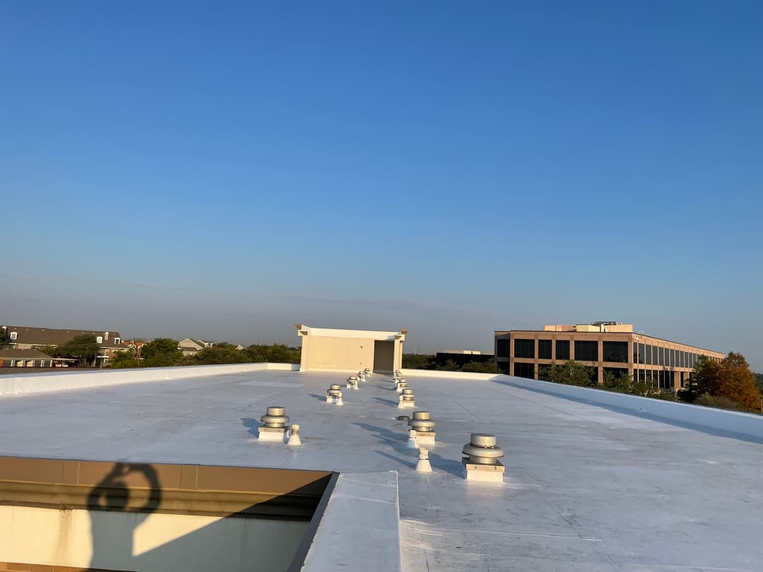 White flat roof with ventilation units under a clear blue sky and a building in the background.