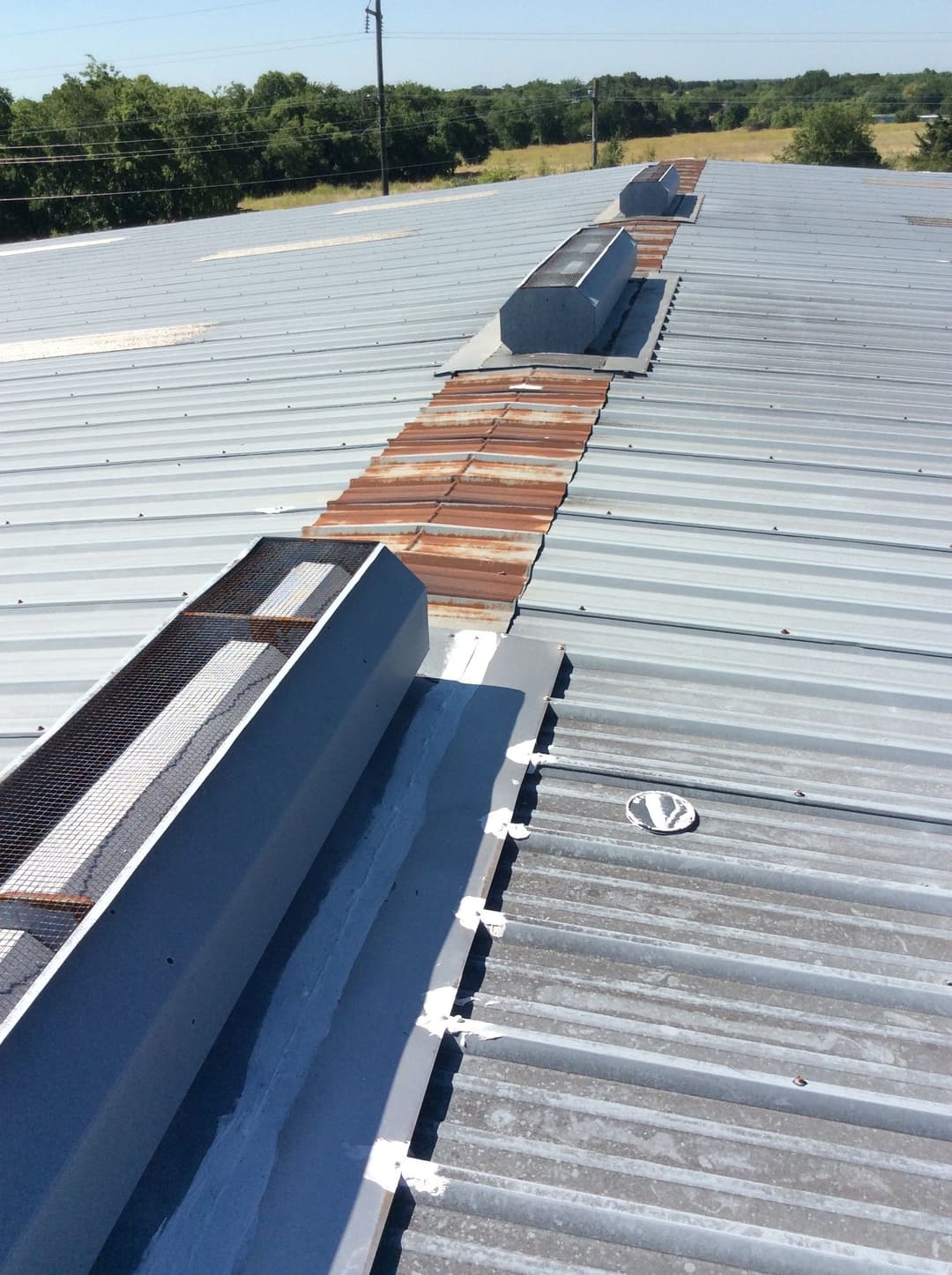 Metal roof with ventilation skylights and patched areas, overlooking green landscape.