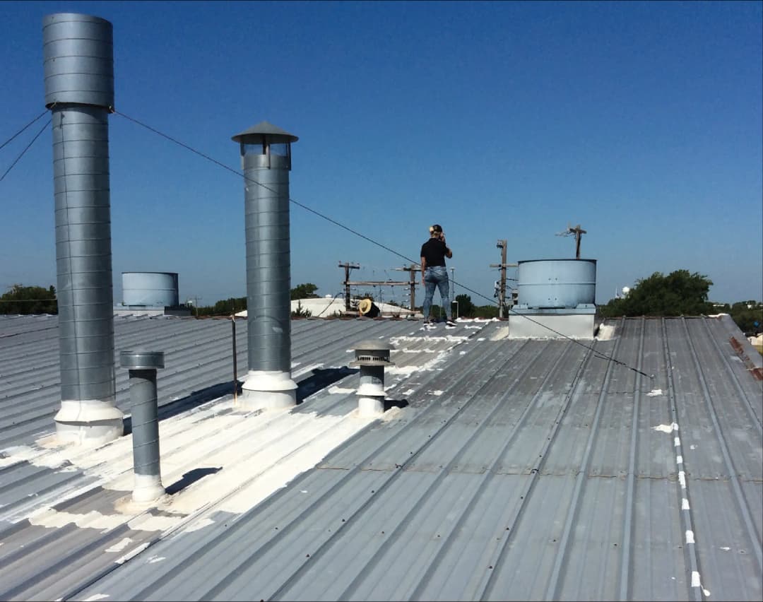 Worker applying coating on a metal roof with ventilation pipes and clear blue sky.