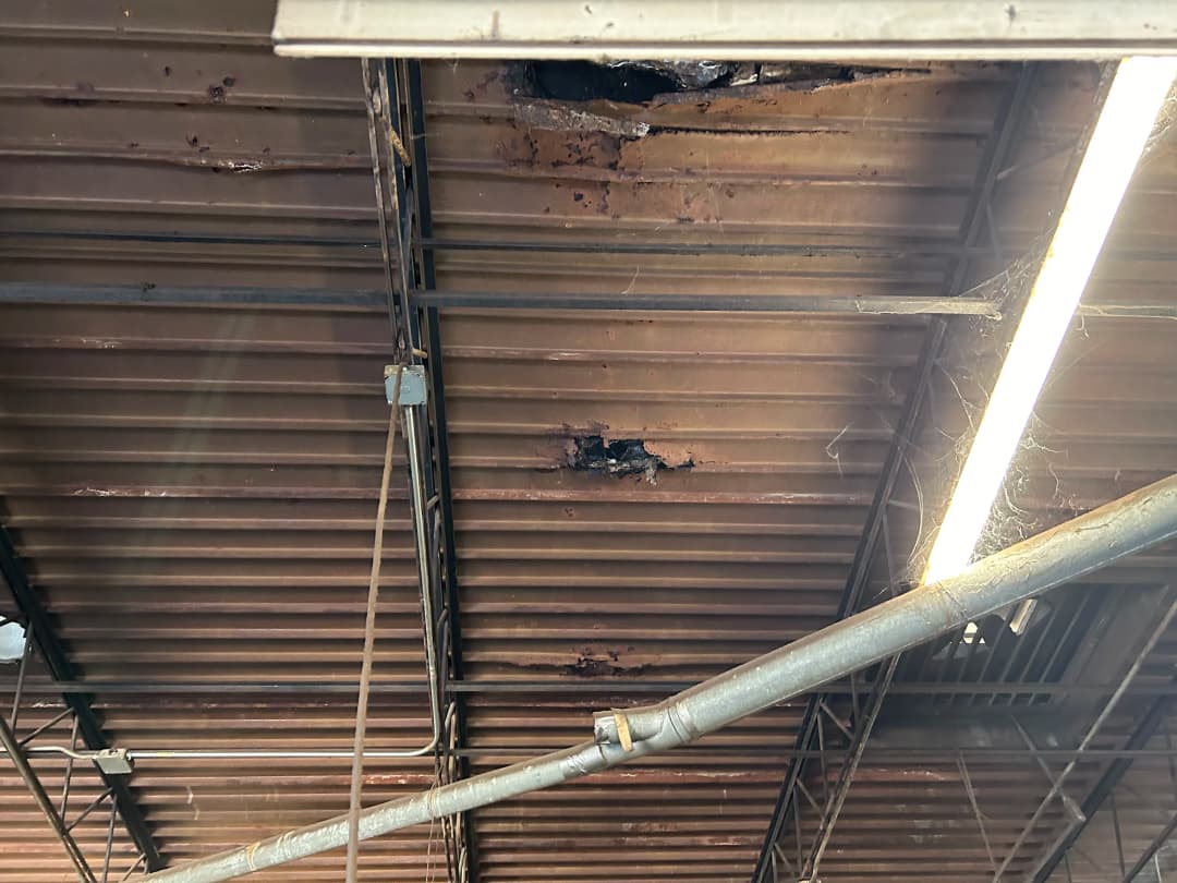 Damaged metal ceiling with rust, holes, and exposed wires in an industrial space.