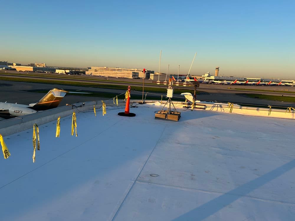 View of airport runway from rooftop, featuring aircraft, safety barriers, and control tower.
