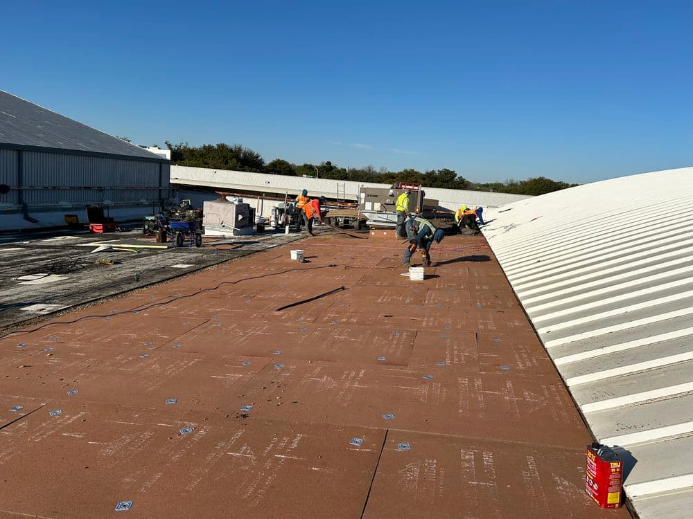 Workers installing roofing materials on a flat commercial roof under a clear blue sky.