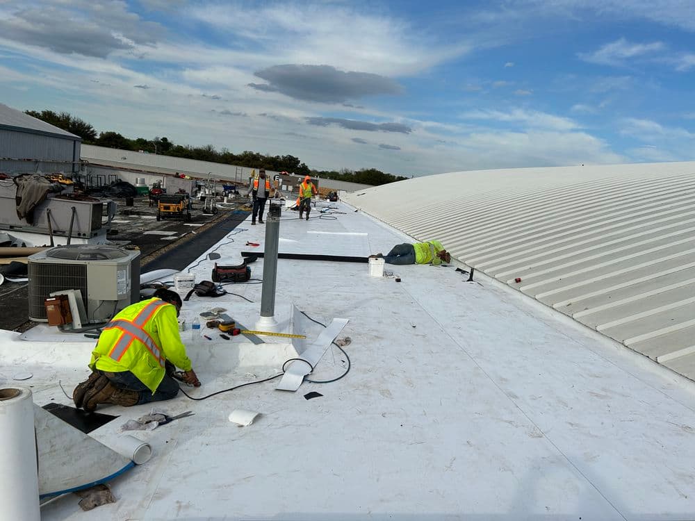Workers installing roofing materials on a commercial building under blue sky and clouds.