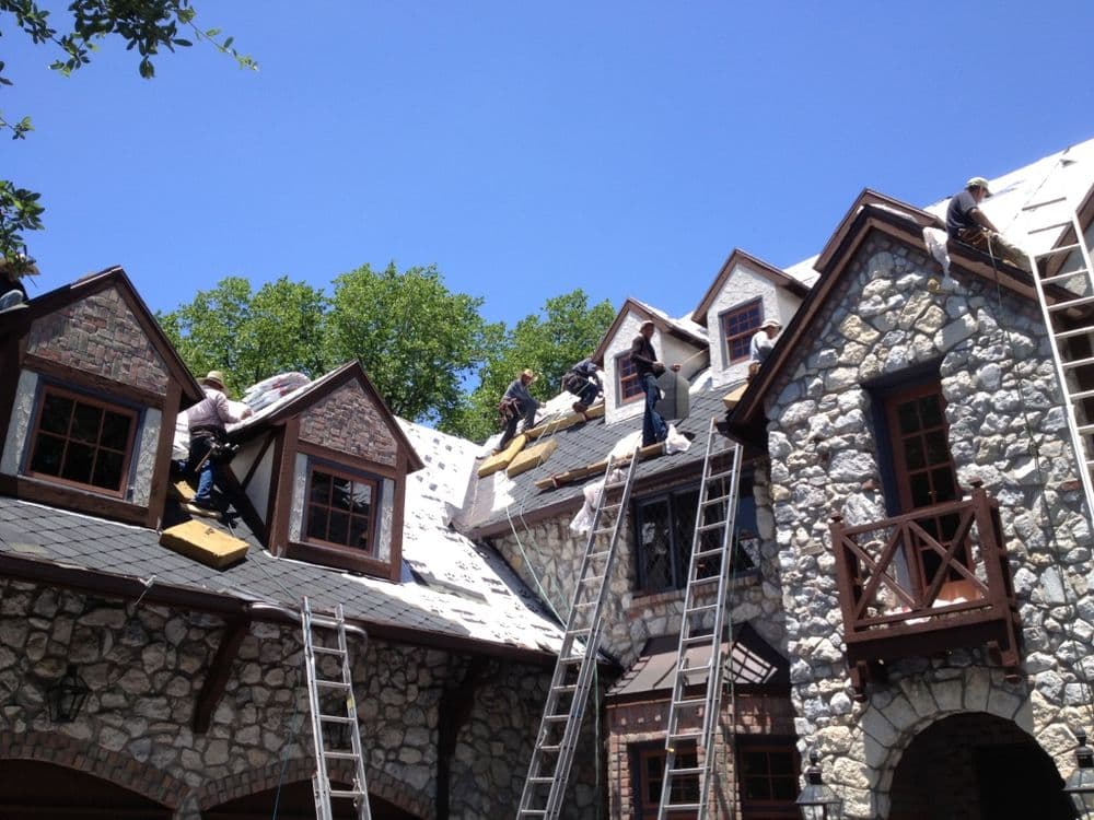 Roofing contractors installing new shingles on a stone house under a clear blue sky.