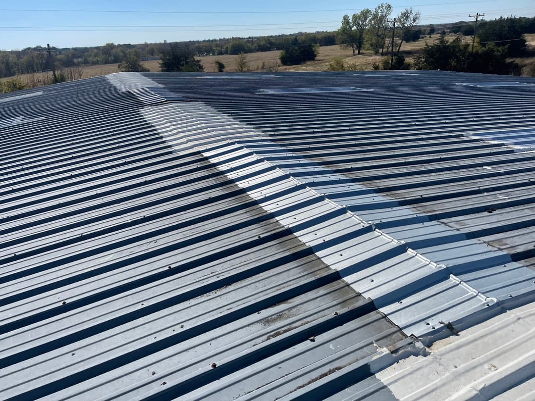 Metal roof with visible seams and patchwork, set against a clear blue sky and grassy landscape.